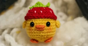 Imagem de um pato amigurumi feito à mão com chapéu de morango, posicionado sobre enchimento branco de fibra. O pato de crochê tem bico e pés laranja, olhos pretos brilhantes e um chapéu vermelho com detalhes verdes e brancos, lembrando uma morango. Ideal para ilustrar a receita completa de Pato Amigurumi com Chapéu de Morango.